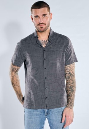 Man with short hair and tattoos on arms and chest wearing a dark gray patterned short-sleeve button-up shirt and light blue jeans.