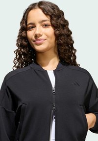 Crni sportski sako s visokim ovratnikom, zatvorenim patentnim zatvaračem sprijeda i bočnim džepovima. Ima akcentu logotipa i mekan, teksturiran materijal.