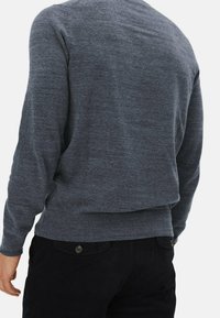 Pull gris en maille avec manches longues, encolure ronde et poignets côtelés. Texture douce et coupe légèrement ample, associé à un pantalon en tissu foncé.