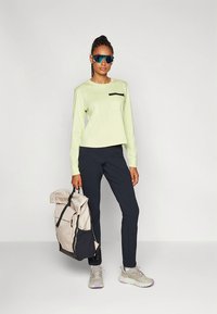 Svetlo zelena majica z dolgimi rokavi s črnim žepnim detajlom, črne hlače, sive superge in bež ter črn nahrbtnik. Sončna očala dokončajo videz.