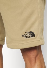 Beige fleece shorts med svart broderad The North Face-logga som bärs på en persons lår.