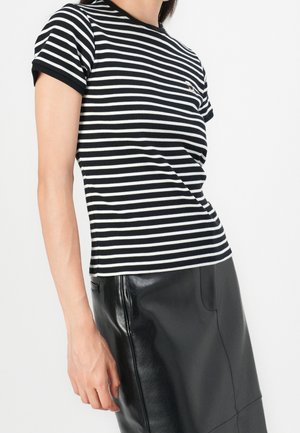 Persoon draagt een zwart-wit gestreept shirt met korte mouwen gecombineerd met een zwarte leren rok met hoge taille, tegen een witte achtergrond.