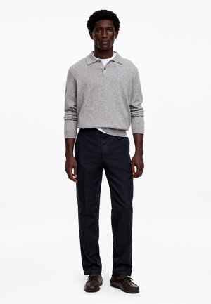 Homme debout portant un polo gris à manches longues, un pantalon foncé et des chaussures habillées marron sur un fond blanc uni.