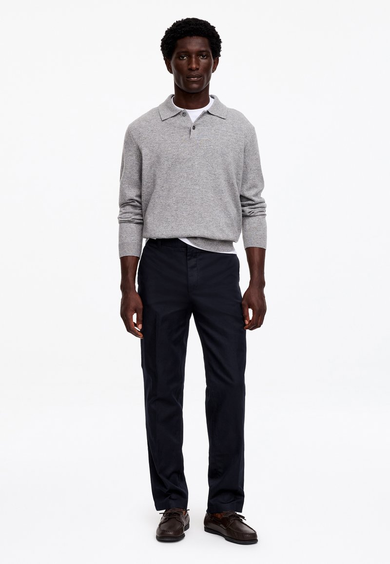 Homme debout portant un polo gris à manches longues, un pantalon foncé et des chaussures habillées marron sur un fond blanc uni.