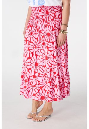 Vrouw draagt een rode en roze midi-rok met bloemen, zilveren sandalen met hakken en gouden armbanden op een witte achtergrond.