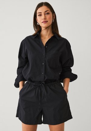 Camisa negra de botones con mangas enrolladas y bolsillo en el pecho, combinada con pantalones cortos negros con cuerda de ajuste, ambos en una tela suave.