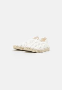 Zapatos blancos tipo slip-on con diseño perforado, suela de goma beige y bordes aserrados. Textura suave y forma redondeada para mayor comodidad.