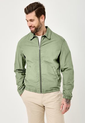 Mann mit Bart, der eine grüne Jacke mit Reißverschluss und beige Hose trägt, Hände in den Taschen, schaut nach unten und lächelt vor einfachem Hintergrund.