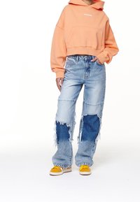 Felpa arancione tagliata con tasca frontale; jeans in denim chiaro e scuro con effetto usurato; sneakers gialle con suola bianca.