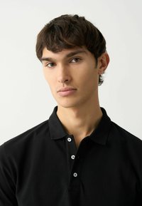 Camisa polo negra con una textura suave, que presenta un collar clásico y tres botones blancos en el cuello, llevada por un modelo.