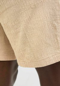 Beige rövidnadrág, strukturált anyagból, gyűrött mintázattal és varrott szegéllyel. Színe lágy és világos, alkalmas mindennapi viseletre.