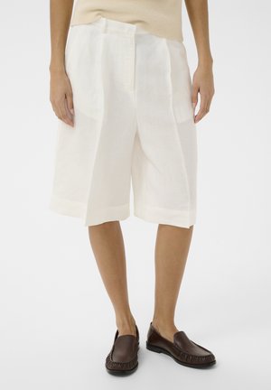 Femme portant un short blanc à jambes larges arrivant aux genoux et des mocassins marron foncé à enfiler, debout devant un fond uni.