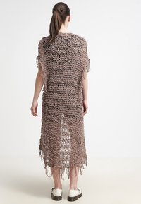 Lång, löst stickad virkade klänning i brunt med fransiga kanter, som har en lös passform och sidosprund för rörelse. Bärs över vita skor.