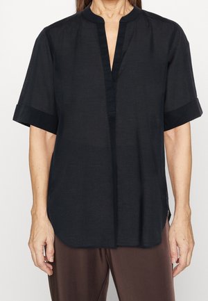 Vrouw die een loszittende zwarte blouse met korte mouwen en een mandarijnkraag draagt, samen met een bruine broek, staand tegen een eenvoudige achtergrond.