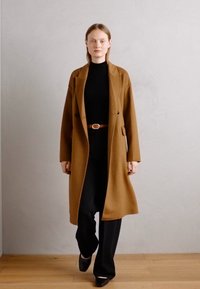 Bruine wollen overjas met een op maat gemaakt ontwerp, voorzien van een riem, gedragen over een zwarte top en wijde broek. Soepele textuur, lengte tot de knie.