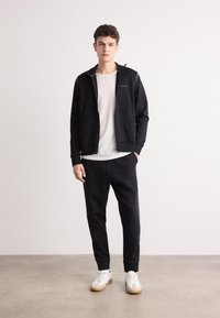 Jachetă neagră cu fermoar, având accente verzi, asortată cu pantaloni jogger negri și adidași albi. Materialul pare moale, cu manșete cu striații și un design ajustat.