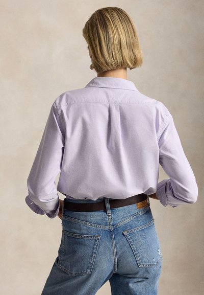Person with short blonde hair wearing a light purple button-up shirt and blue jeans, shown from the back against a plain background.