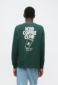 Zelena majica dugih rukava s grafičkim printom na leđima koji sadrži tekst "ICED COFFEE CLUB" i ilustraciju šalice.