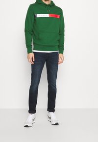 Mężczyzna w zielonej bluzie z kapturem Tommy Hilfiger, ciemnoniebieskich dżinsach i białych butach sportowych Nike stoi na tle jednolitego białego tła.