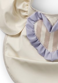 Cremefarvet forklæde med volang i lavendel. I midten er der et hjerteformet patch med lodrette beige og creme striber. Glat tekstur hele vejen igennem.