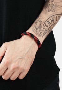 Braccialetto intrecciato nero e rosso sul polso; presenta un design testurizzato e intrecciato. La mano è parzialmente visibile su uno sfondo di camicia nera.
