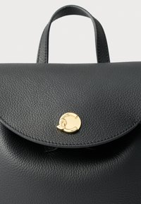 Mochila em pele preta com superfície texturada, aba dobrável e fecho circular dourado. Apresenta alças curtas na parte superior para transporte.