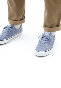 Baskets en daim bleu avec semelles en caoutchouc blanches et lignes d'accent noires, associées à un pantalon kaki retroussé. Les baskets présentent un design à lacets et des détails de couture.