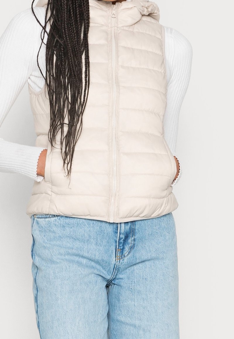 Gilet imbottito beige con cappuccio, caratterizzato da una cerniera frontale, design trapuntato e tasche laterali, indossato sopra una maglietta a maniche lunghe bianca.