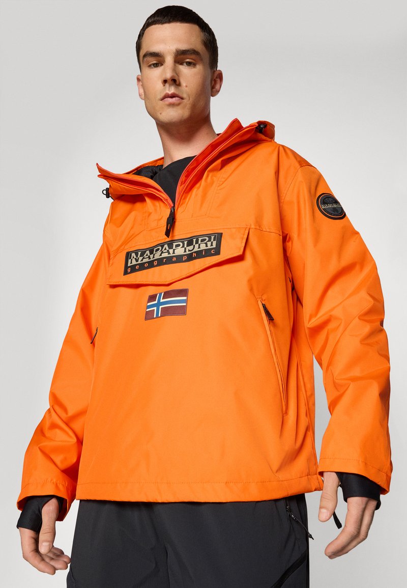 Giacca impermeabile arancione con tasca frontale con zip, patch con logo e zip laterali. Presenta un colletto alto e polsini elasticizzati.