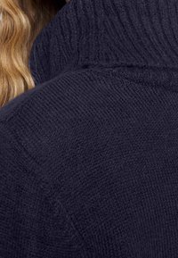 Cheveux blonds partiellement visibles derrière une personne portant un pull épais en tricot bleu foncé avec un col montant.