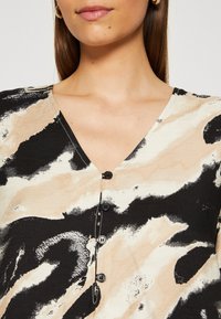 Knappet skjorte med V-hals, der har et abstrakt tryk i sort, beige og hvid. Blød tekstur med sorte knapper foran.