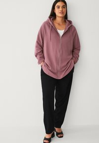 Stofroze hoodie met rits en capuchon, ruime pasvorm, voorzakken, gecombineerd met losse zwarte broek en zwarte open-toe hakken. Gladde textuur.