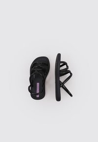 Sandalias negras con múltiples correas entrelazadas y una plantilla texturizada. La plantilla tiene una etiqueta púrpura que muestra el nombre de la marca.