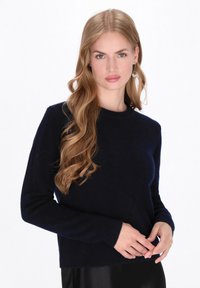 Vrouw met lang golvend blond haar, gekleed in een marineblauwe trui en hangende oorbellen, die haar mouwen aanpast tegen een witte achtergrond.
