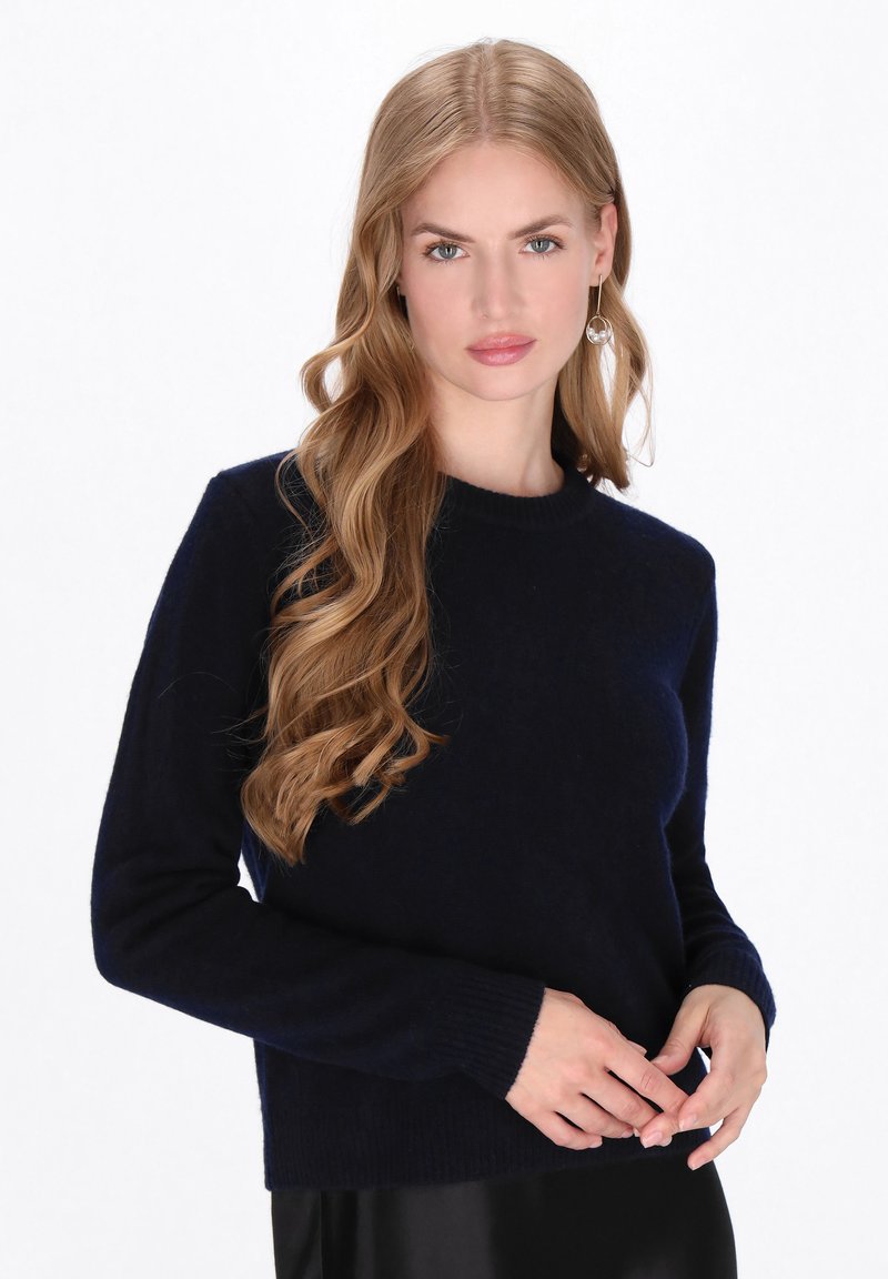 Vrouw met lang golvend blond haar, gekleed in een marineblauwe trui en hangende oorbellen, die haar mouwen aanpast tegen een witte achtergrond.