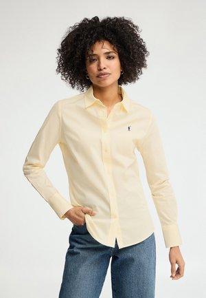 Donna con capelli ricci che indossa una camicia giallo pallido abbottonata e jeans blu, in piedi con una mano in tasca contro uno sfondo neutro.