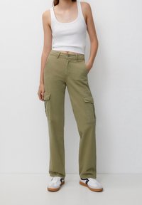 Femme portant un pantalon cargo vert olive, un débardeur blanc et des baskets blanches à rayures noires, debout devant un fond clair uni.