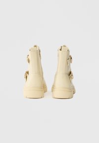 Botas de tornozelo em cor creme com acabamento brilhante, apresentando duas correias com fivela e um fecho lateral. Sola de borracha texturizada para melhor aderência.