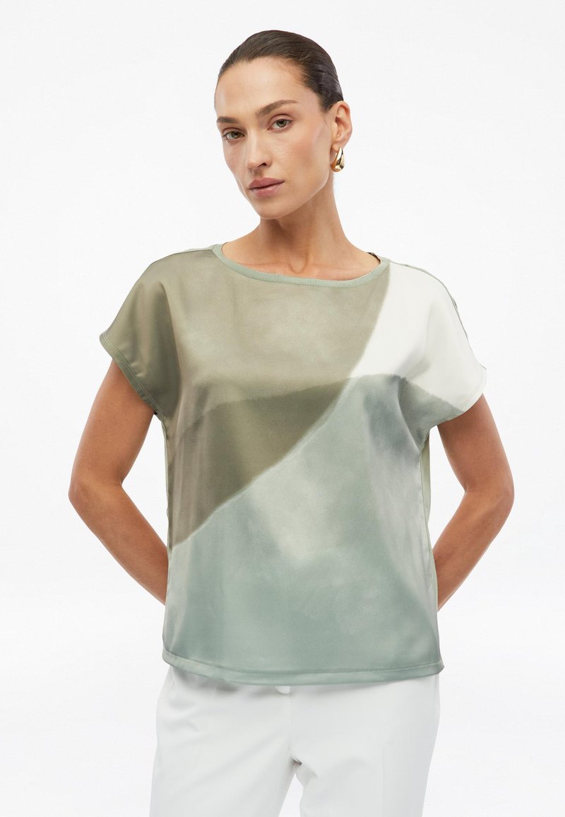 Woman with dark hair wearing gold hoop earrings and a short-sleeve, abstract green and white color-blocked top with white pants.