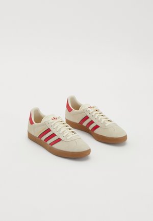 Paire de baskets en daim beige avec des rayures rouges et un détail au talon, semelles en gomme et lacets crème sur fond blanc.