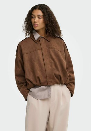 Frau mit lockigem Haar, die eine braune Wildlederjacke über einem gestreiften Hemd und hellbeigen Hosen trägt, Hände in den Taschen, schaut nach links.