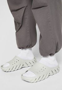 Ljusgrå slip-on sandaler med öppen tå, texturerade hål på ovansidan och tjock, vågig sula. Bärs med vita strumpor och grå joggerbyxor.