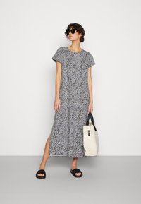 Vestido maxi em padrão de folhas em preto e branco, mangas curtas, com fenda lateral, feito de tecido leve, combinado com sandálias pretas e uma bolsa bege.