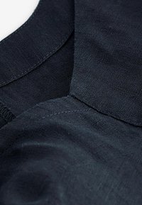 Tessuto blu scuro che mette in evidenza una trama di lino testurizzato. Presenta un colletto arrotondato e una cucitura evidente lungo i bordi.
