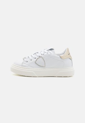 White leather sneaker with stitched detailing, round toe, and beige metallic accent on the heel. Features flat white laces and textured sole.