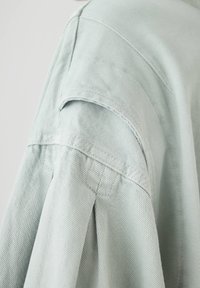 Chemise en tissu texturé vert clair avec un pli d'épaule et des détails de poignetulés. Présente des coutures plates et un design de col classique.