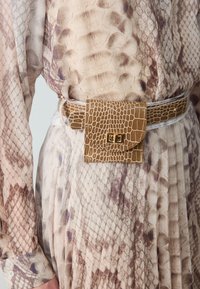 Cintura in pelle effetto coccodrillo color marrone chiaro con una tasca rettangolare e chiusura in tonalità oro, indossata sopra un vestito in stampa serpente testurizzata nei toni beige e viola.