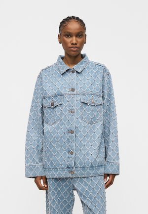 Femme portant une veste et un pantalon en denim bleu clair avec un motif en losanges cousu et de petites embellissements ressemblant à des perles.
