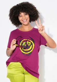 Magenta t-shirt med et sort og neongult grafisk motiv af et kranium med et smilende ansigt. Båret med limegrønne bukser, model der holder solbriller.