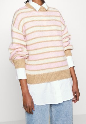 Persoon draagt een gebreide trui met roze, witte en beige strepen over een wit overhemd met kraag en lichtblauwe spijkerbroek.